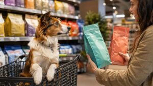 Perro en un carrito observando cómo su propietaria compara dos alimentos para mascotas en el punto de venta.